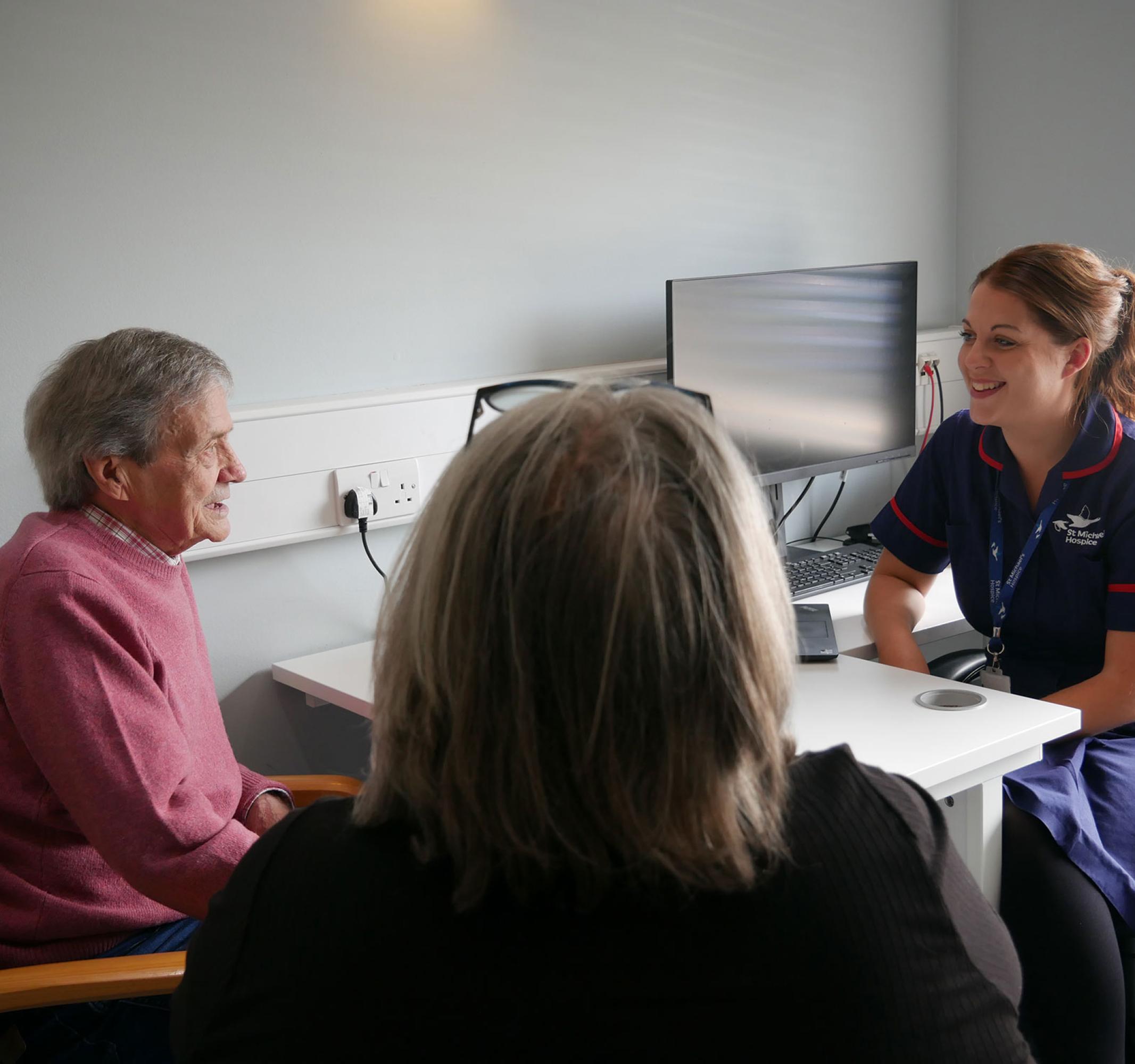 Nurse smiles, talking with two patients in a consultation room.