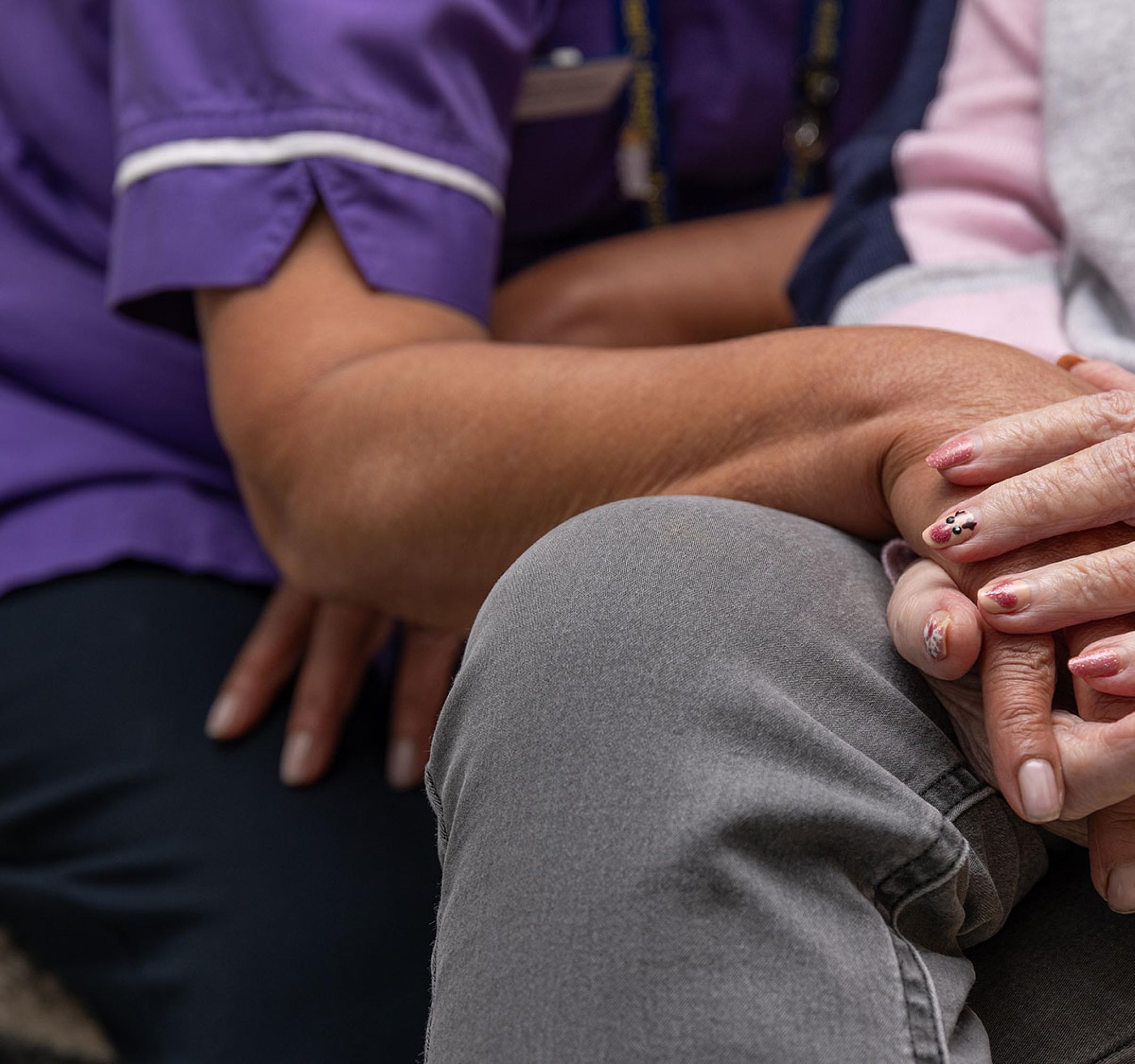 A person in a purple uniform gently holds the hand of an elderly person.