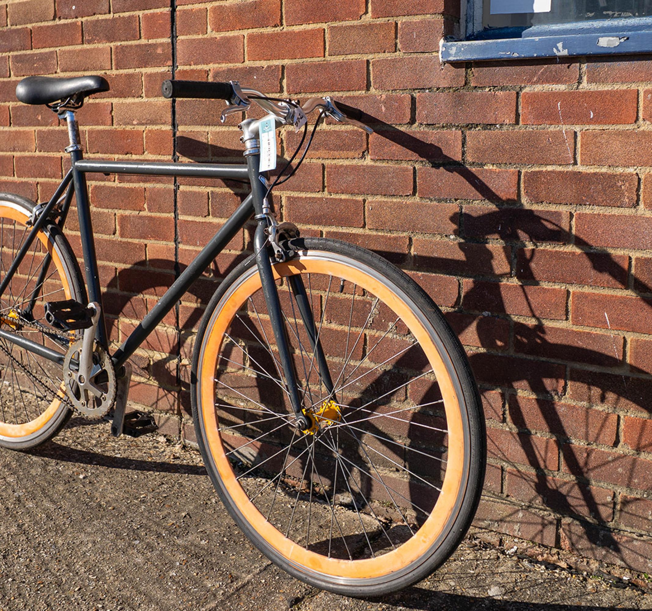 Black bicycle with orange wheels leans against a brick wall, sunlight creating a long shadow.