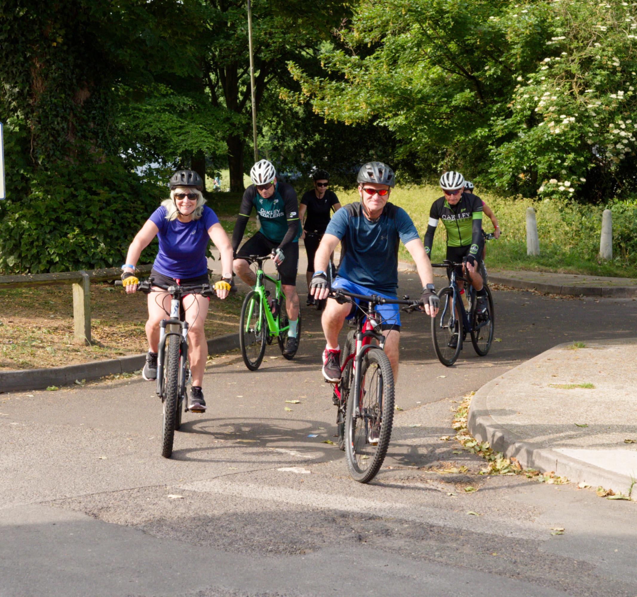 Five cyclists ride together on a paved path, trees and a parking sign in the background.