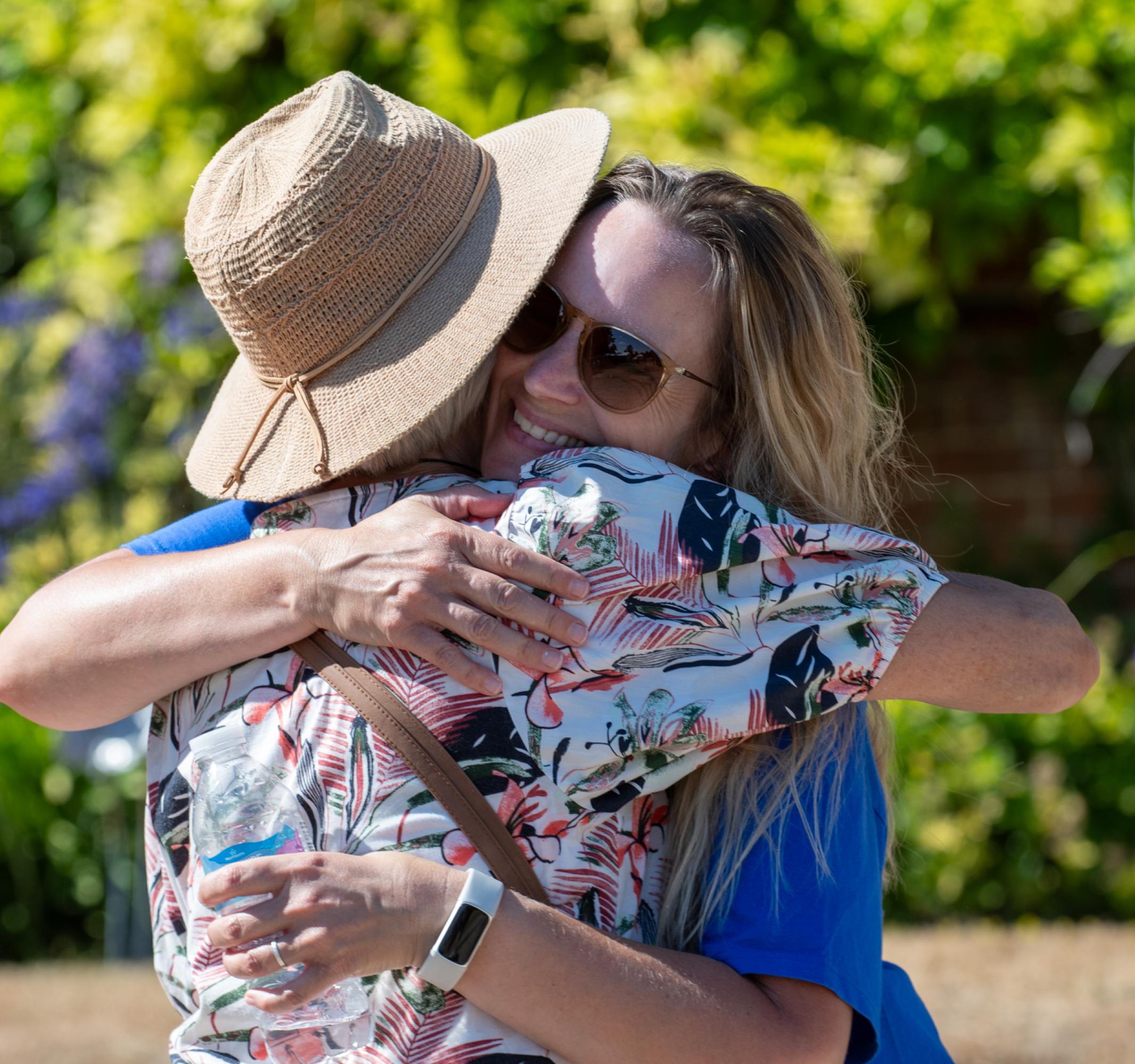 Two people sharing a warm hug outdoors, one in a hat, the other in sunglasses.