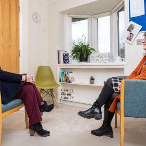Two people sit in chairs facing each other in a bright room with a bay window.