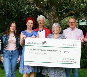 Photograph: Eight people holding a large check for The Camphill Trust in a green outdoor setting.