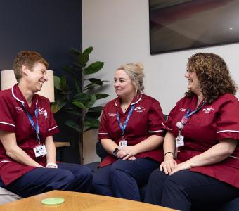 Three smiling women in healthcare uniforms chat on a blue sofa.