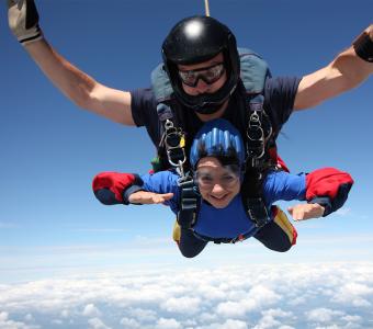 Two people tandem skydiving against a bright blue sky, smiling.