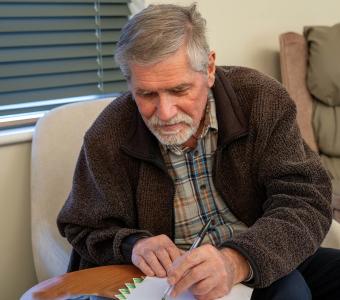 Grey-haired man in a brown jacket writing intently.