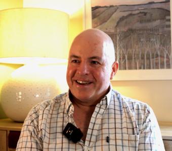 Man in plaid shirt smiles, wearing a lapel mic. Warm room with lamp and artwork.