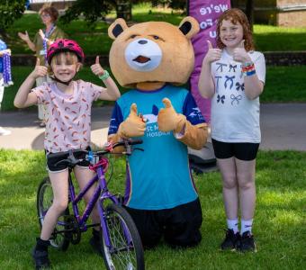 Two girls and a bear mascot give thumbs up with a purple bike on green grass.