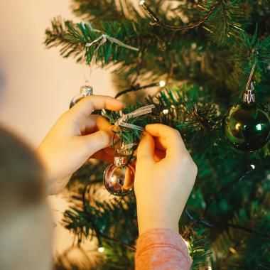 Child's hands hanging a silver ornament on a green Christmas tree with lights.
