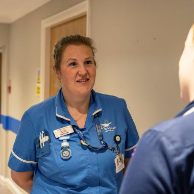 Smiling nurse in blue scrubs talks to colleague in a hospital hallway.