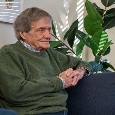 Man in green sweater sitting on sofa, looking right with clasped hands.