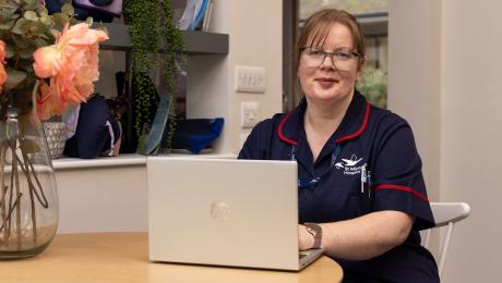 Smiling nurse in blue uniform working on a laptop at a wooden table.