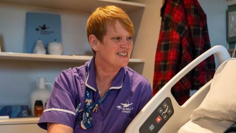 Friendly nurse in purple uniform talks to a patient in a hospital bed.