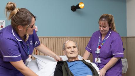 Two nurses assist an elderly male patient sitting up in a hospital bed.