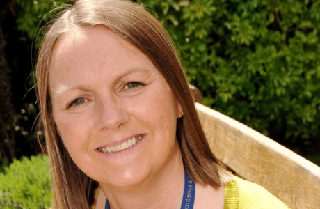Smiling woman with long brown hair, yellow top, and blue lanyard outdoors.