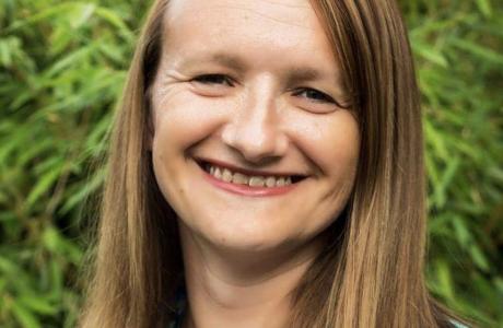 Smiling woman with long brown hair, wearing a blue top, against a green background.