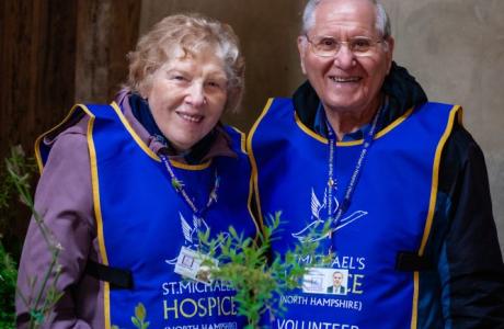 Smiling senior man and woman in blue hospice volunteer vests stand behind potted plants.