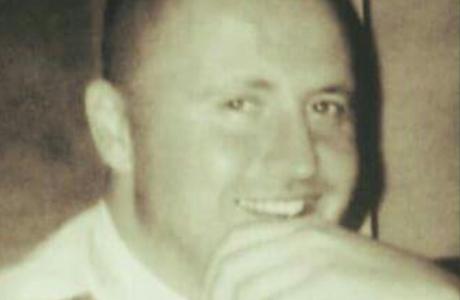 Sepia photograph: Smiling man with shaved head, hand to chin.