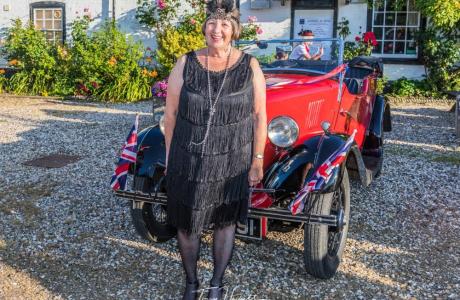 Smiling woman in flapper dress stands by a red vintage car with Union Jack flags.