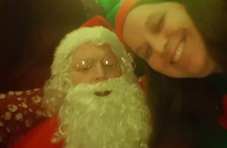 Santa Claus and a smiling woman in an elf hat under colorful festive lights.
