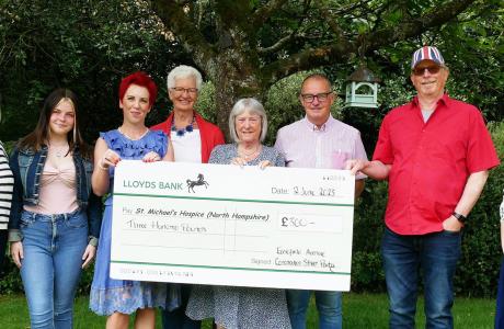 Photograph: Eight people holding a large check for The Camphill Trust in a green outdoor setting.