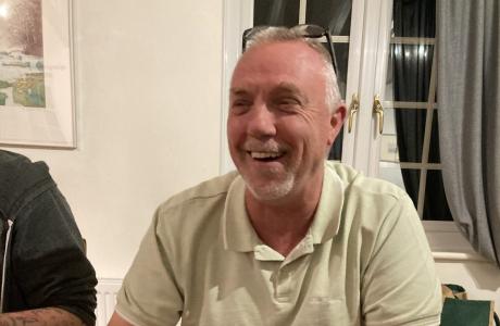 Middle-aged man with grey hair and beard laughing heartily indoors.