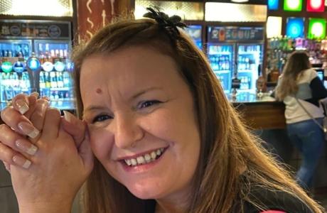 Smiling woman with long hair and clasped hands in a pub, bottles and taps in background.
