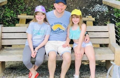 Smiling man and two young girls in hats sitting on a wooden bench outdoors.
