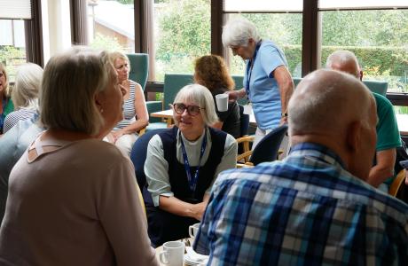People socialising, some holding mugs, in a bright room with large windows.