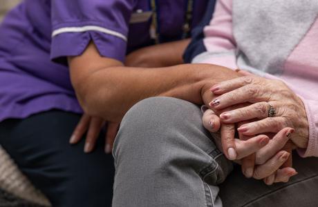 A person in a purple uniform gently holds the hand of an elderly person.