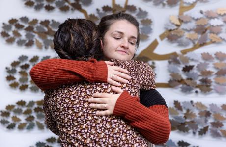 Two women hugging warmly in front of a metallic leaf wall art.