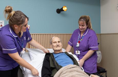 Two nurses assist an elderly man sitting up in a hospital bed, smiling.