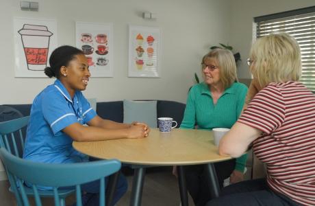 Healthcare professional in blue scrubs talks with two women at a table.
