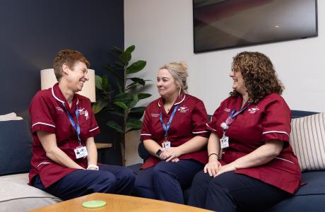 Photograph: Three healthcare professionals in maroon scrubs smiling and conversing on a blue couch.