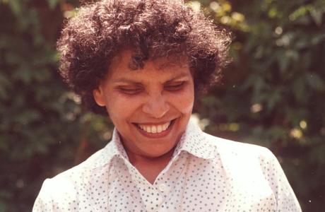 Smiling woman with curly hair holding a light pink rose in a garden.