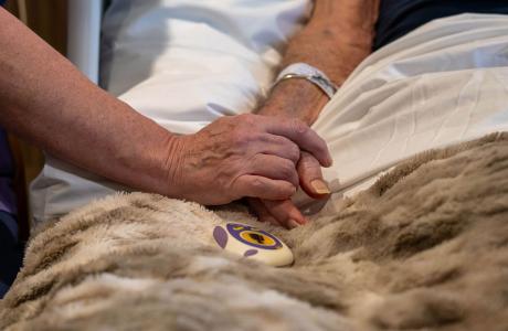 A comforting hand gently holds a frail hand with a hospital ID band on a fuzzy blanket.