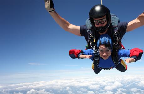 Two people tandem skydiving against a bright blue sky, smiling.