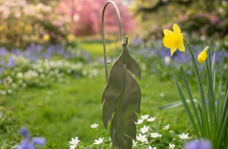 Metal leaf garden decoration among blue, white, and yellow flowers in a sunny spring garden.