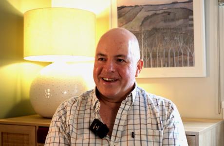 Man in plaid shirt smiles, wearing a lapel mic. Warm room with lamp and artwork.