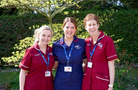Three smiling healthcare professionals in uniforms, one blue and two burgundy, outdoors.