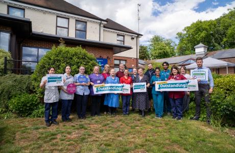 Group of smiling healthcare staff holding "Outstanding" signs in front of a care facility.