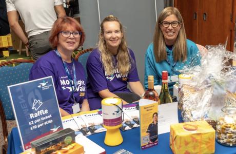 Smiling women at an event table with a raffle sign, collection bucket, and prizes.