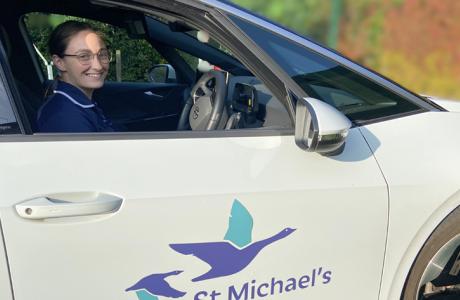 Woman in uniform smiles from white car with St Michael's hospice bird logo.