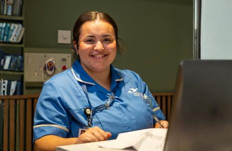 Smiling nurse in blue scrubs at a hospital desk, looking at the camera.