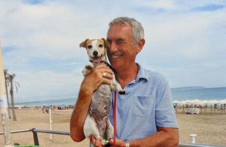 Grey-haired man smiles, holding a small white and tan dog on a beach boardwalk.