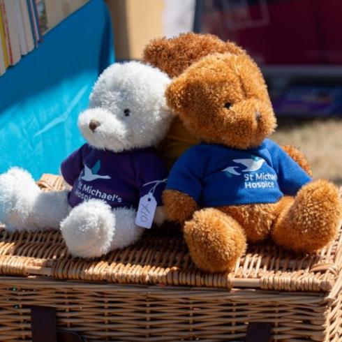White and brown teddy bears, wearing St Michael's Hospice shirts, sit on a wicker basket.