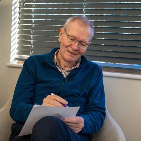 Happy older man in glasses and blue fleece writes on paper, seated in a chair.