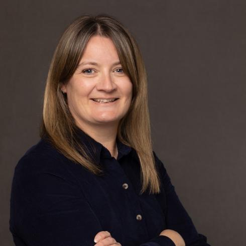 Smiling woman with light brown hair, wearing a dark blue top, arms crossed.