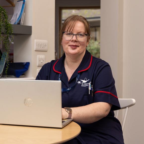 Smiling nurse in blue uniform working on a laptop at a wooden table.