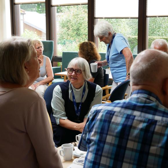 People socialising, some holding mugs, in a bright room with large windows.